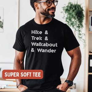 K&ouml;nnte beinhalten: Schwarzes T-Shirt mit wei&szlig;em Text: "Hike & Trek & Walkabout & Wander." Ein rotes Rechteck unten lautet "SUPER SOFT TEE." Das Shirt wird von einer Person mit Brille und Bart getragen.