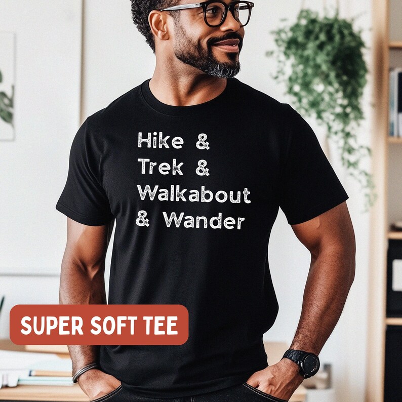 K&ouml;nnte beinhalten: Schwarzes T-Shirt mit wei&szlig;em Text: "Hike & Trek & Walkabout & Wander." Ein rotes Rechteck unten lautet "SUPER SOFT TEE." Das Shirt wird von einer Person mit Brille und Bart getragen.