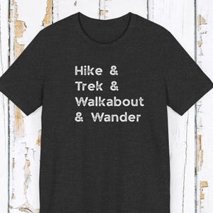K&ouml;nnte beinhalten: Dunkelgraues T-Shirt mit den wei&szlig;en Worten "Hike & Trek & Walkabout & Wander". Das Shirt hat einen Rundhalsausschnitt und kurze &Auml;rmel. Der Text ist vertikal gestapelt.