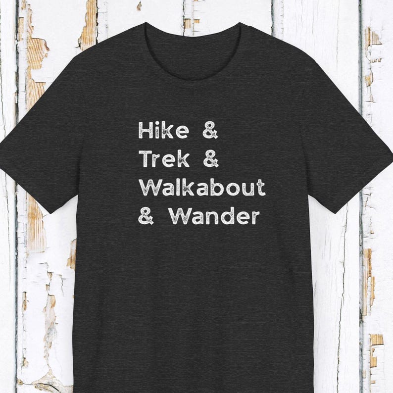 K&ouml;nnte beinhalten: Dunkelgraues T-Shirt mit den wei&szlig;en Worten "Hike & Trek & Walkabout & Wander". Das Shirt hat einen Rundhalsausschnitt und kurze &Auml;rmel. Der Text ist vertikal gestapelt.