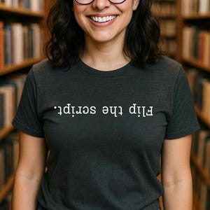 May include: Dark gray t-shirt with the text "Flip the script" in white. The shirt is a crew neck style and is worn by a person with glasses and dark hair. The background is a library with bookshelves.