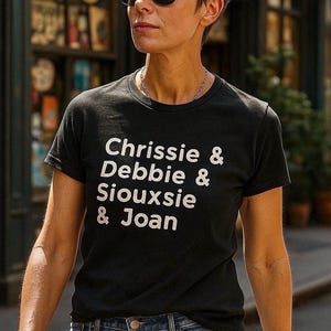 Op de afbeelding: Zwart T-shirt met de namen "Chrissie & Debbie & Siouxsie & Joan" in witte letters. De persoon draagt een zonnebril en een blauwe spijkerbroek. De achtergrond is wazig, wat wijst op een buitenomgeving.