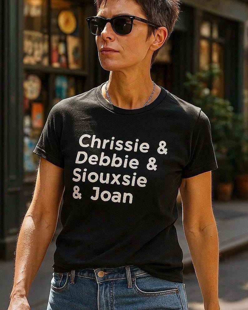 Op de afbeelding: Zwart T-shirt met de namen "Chrissie & Debbie & Siouxsie & Joan" in witte letters. De persoon draagt een zonnebril en een blauwe spijkerbroek. De achtergrond is wazig, wat wijst op een buitenomgeving.