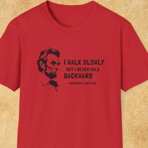 May include: Red t-shirt featuring a black silhouette of Abraham Lincoln and the quote, "I walk slowly but I never walk backward - Abraham Lincoln." The shirt has a crew neck and short sleeves.