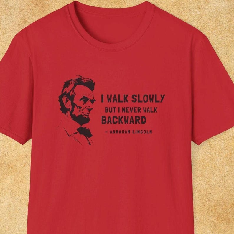 May include: Red t-shirt featuring a black silhouette of Abraham Lincoln and the quote, "I walk slowly but I never walk backward - Abraham Lincoln." The shirt has a crew neck and short sleeves.