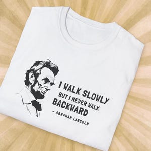 May include: White t-shirt featuring a black and white illustration of Abraham Lincoln. The shirt includes the quote "I walk slowly but I never walk backward" attributed to Abraham Lincoln.