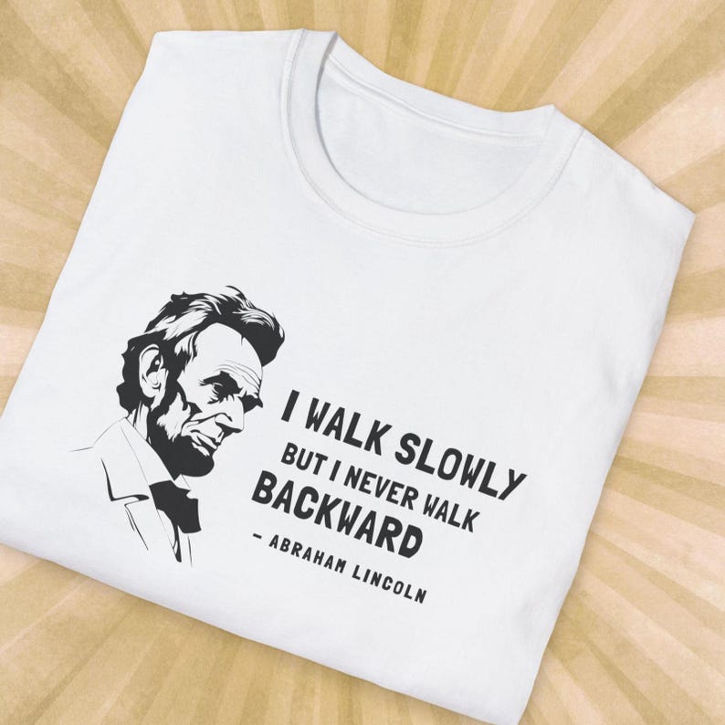May include: White t-shirt featuring a black and white illustration of Abraham Lincoln. The shirt includes the quote "I walk slowly but I never walk backward" attributed to Abraham Lincoln.
