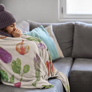 May include: A person wrapped in a beige blanket adorned with watercolor illustrations of vegetables, including eggplant, onions, and asparagus. The person is wearing a purple beanie and scarf, sitting on a gray couch.