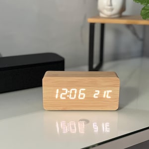 Könnte beinhalten: Eine rechteckige digitale Holzuhr, die die Uhrzeit 12:06 und das Datum 21 anzeigt. Die Uhr hat eine helle Holzmaserung. Die Uhr steht auf einer weißen Oberfläche mit einer schwarzen Soundbar und einer Buddha-Kopf-Statue im Hintergrund.