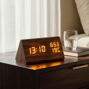 May include: A triangular dark wood digital alarm clock with an orange LED display showing the time 13:10, a humidity level of 65% RH, and a temperature of 19 degrees Celsius, resting on a dark wooden nightstand next to a glass of water.