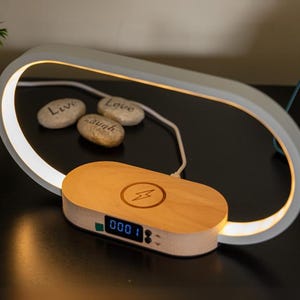 Peut inclure: Station de recharge sans fil moderne avec une base en bois et un écran numérique. La station est éclairée par une lumière blanche de forme ovale. Trois petites pierres avec les mots "Live, Love, Laugh" sont à proximité.