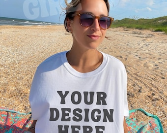 May include: A white t-shirt with the text "YOUR DESIGN HERE" in a bold, dark grey font. The person in the image is wearing sunglasses and is on a beach. The background includes the ocean, sand, and a blue sky.