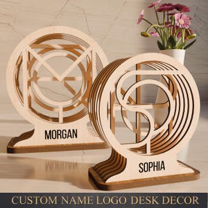 Op de afbeelding: Twee houten bureau-decoraties met aangepaste naamlogo's. Elk stuk heeft een cirkelvormig ontwerp met gelaagde, geometrische uitsparingen. De namen "MORGAN" en "SOPHIA" worden op de basis weergegeven. De onderkant van de afbeelding luidt "CUSTOM NAME LOGO DESK DECOR".