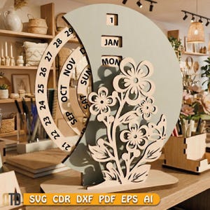 May include: A wooden perpetual calendar with a light green crescent-shaped background and a floral design. The calendar displays the month, day, and date. The base is made of light-colored wood. The text "JAN" and "MON" are visible.