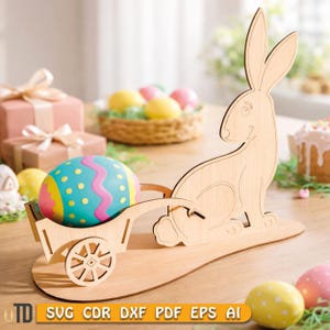 Könnte beinhalten: Osterdekoration aus Holz mit einem Hasen und einem Wagen, der ein dekoriertes Ei trägt. Der Hase ist hellbraun und sitzt auf einem Holzsockel. Der Wagen hat ein großes, buntes Ei mit blauen, rosa und gelben Akzenten. Der Text "SVG CDR DXF PDF EPS AI" ist enthalten.