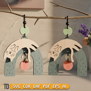 Może przedstawiać: Drewniane kolczyki w kształcie kota z jasnym wykończeniem drewna. Kolczyki mają wzór śpiącego kota nad niebiesko-zielonym łukiem, z różowym kółkiem i zielonym kółkiem. Kolczyki wiszą na czarnym haczyku.