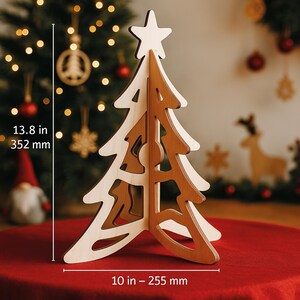 May include: A wooden Christmas tree decoration with a star on top. The tree is made of light and dark wood, measuring 13.8 inches (352 mm) tall and 10 inches (255 mm) wide. The tree is on a red surface, with Christmas decorations in the background.