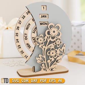 May include: A wooden perpetual calendar with a light gray crescent moon shape and a wooden base. The calendar displays the date, month, and day. A laser-cut floral design is on the front. The calendar is approximately 7 inches tall.