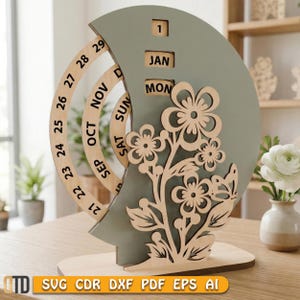 May include: A wooden perpetual calendar with a sage green crescent moon shape and a floral design. The calendar displays the month, day, and date. The base is made of light-colored wood. The text "SVG CDR DXF PDF EPS AI" is at the bottom.