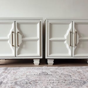 May include: Two white bedside cabinets with gold-coloured handles and trim. Each cabinet has two doors with a decorative panel design. The cabinets are on short, square legs and sit on a patterned rug.