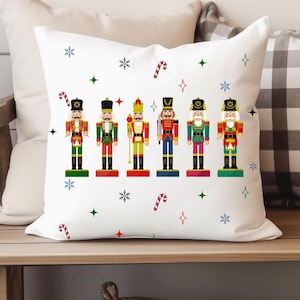 Puede incluir: Cojín decorativo blanco con una fila de soldados cascanueces de colores. El cojín tiene copos de nieve, bastones de caramelo y detalles de estrellas. El cojín está sobre un banco de madera con otros cojines y un pequeño árbol de Navidad.