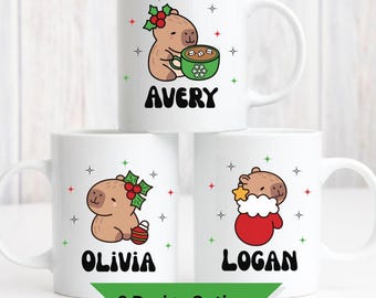 Può includere: Tazze in ceramica bianca con disegni di capibara dei cartoni animati. Una tazza presenta un capibara con una bacca di agrifoglio, un'altra con una tazza di cacao e un'altra con una stella in un guanto. I nomi Avery, Olivia e Logan sono stampati sulle tazze.