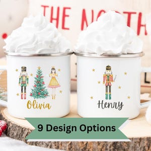 May include: Two white enamel mugs with whipped cream, featuring Christmas-themed designs. One mug has a nutcracker, Christmas tree, and ballerina with the name "Olivia." The other mug has a nutcracker and the name "Henry." The mugs are on a wooden surface.
