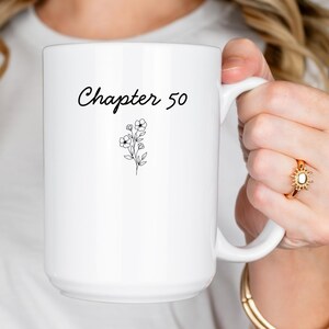 以下が含まれることがあります： 白いセラミックマグカップ。筆記体の「Chapter 50」の文字と小さな花のイラストが描かれています。マグカップには大きなハンドルが付いています。
