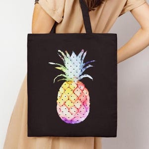Può includere: Borsa tote nera con un colorato design ad acquerello di ananas. L'ananas è raffigurato in tonalità di giallo, rosa, blu e viola, con un motivo a pois. La borsa ha manici neri e viene portata a spalla.