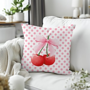 Puede incluir: Un cojín decorativo con un diseño de acuarela de dos cerezas rojas y un lazo rosa. El cojín tiene un fondo blanco con un patrón de lunares rosas. El cojín está sobre un sofá blanco.