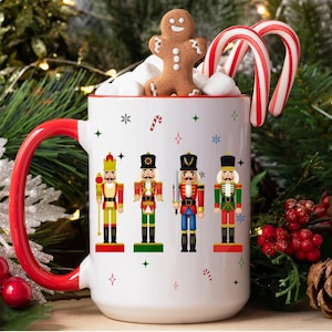 May include: A white ceramic mug with a red handle, decorated with four nutcracker figures. The mug is filled with marshmallows, a gingerbread man, and two candy canes. Festive holiday theme.