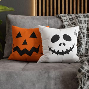 May include: Two Halloween-themed throw pillows on a gray couch. One pillow is orange with a black jack-o'-lantern face. The other is white with a black ghost face and a spider. A plaid blanket is draped over the couch.