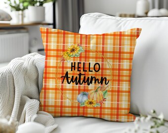 May include: Square cushion with an orange and white check pattern. The cushion features the text "HELLO Autumn" inside a gold wreath, with illustrations of sunflowers and pumpkins. The cushion is on a white sofa.