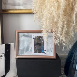 May include: A framed playing card, the three of clubs, with abstract art in a rose gold frame. The frame sits on a black box, with a white shopping bag and a blue vase in the background. Pampas grass hangs from above.