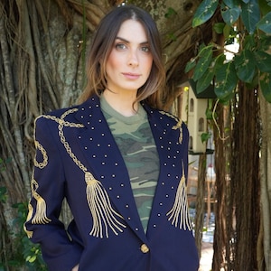 May include: A navy blue blazer with gold chain and tassel embroidery. The blazer has a single button closure and is worn over a camouflage print t-shirt.