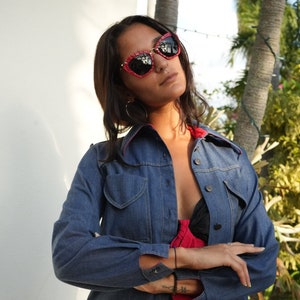May include: A woman wearing a blue denim shirt with a red trim, a red and black patterned skirt, and red cat-eye sunglasses.