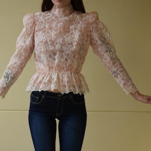 May include: A woman wearing a light pink lace blouse with a high neck and long sleeves. She is also wearing dark blue jeans with embroidered details on the legs.