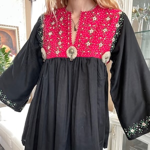 May include: Black tunic top with a red yoke adorned with white floral embroidery. The yoke features a V-neck and is embellished with silver-toned metal accents. The long sleeves and hem are also decorated with floral embroidery. The top is paired with blue jeans.