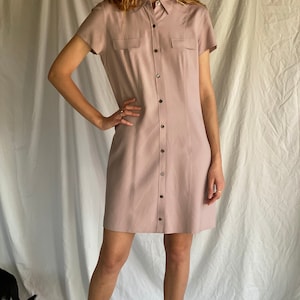 Puede incluir: Un vestido camisa de color rosa claro con botones, mangas cortas y cuello. El vestido tiene una silueta ajustada y llega a la mitad del muslo. La modelo lleva el vestido con botines marrones.