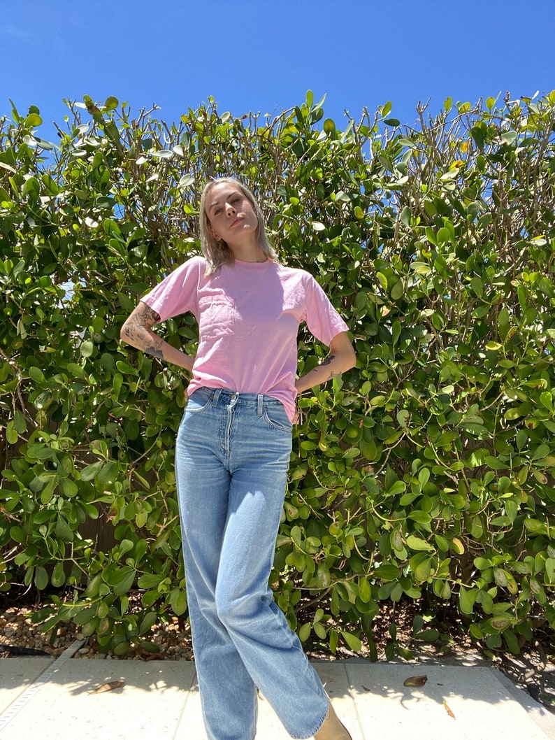May include: A person wearing a light pink t-shirt and light blue jeans standing in front of a green bush.