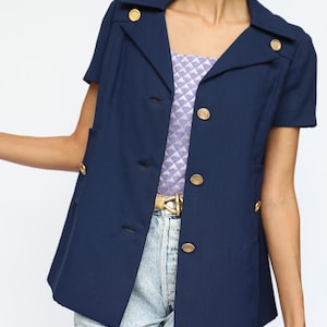 May include: A navy blue blazer with short sleeves and gold buttons. The blazer has a collar and a single breasted closure. The blazer is worn over a purple and white patterned top and light blue jeans.