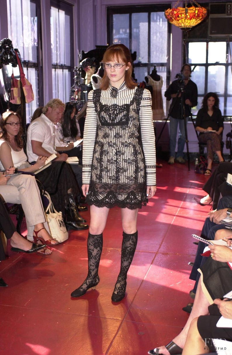 Pode incluir: Uma modelo em um vestido listrado preto e branco com uma sobreposi&ccedil;&atilde;o de renda preta e uma bainha de renda combinando. O vestido &eacute; combinado com meias de renda pretas na altura do joelho e sapatos pretos. A modelo est&aacute; usando &oacute;culos.