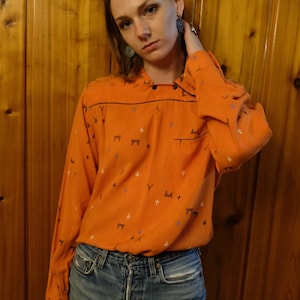 Puede incluir: Una mujer con una camisa de manga larga de color naranja brillante con un patrón geométrico y pantalones vaqueros azules. La camisa tiene cuello y cierre de botón.