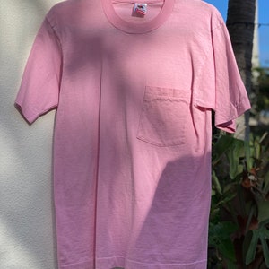 May include: A pale pink t-shirt with a single pocket on the left side. The shirt is hanging on a black hanger against a white wall.