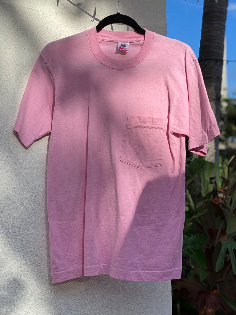 May include: A pale pink t-shirt with a single pocket on the left side. The shirt is hanging on a black hanger against a white wall.