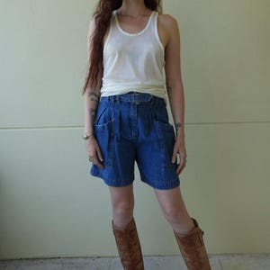 May include: A white sleeveless tank top paired with high-waisted blue denim shorts featuring a matching fabric belt with a buckle, large side pockets, and pleated detailing at the waist, worn with knee-high brown leather boots.