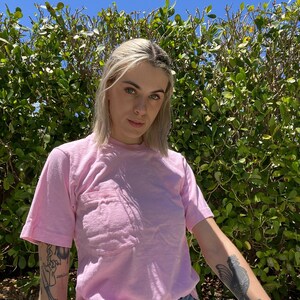 May include: A woman wearing a pink t-shirt with a pocket and blue jeans. She has short blonde hair and tattoos on her arms.