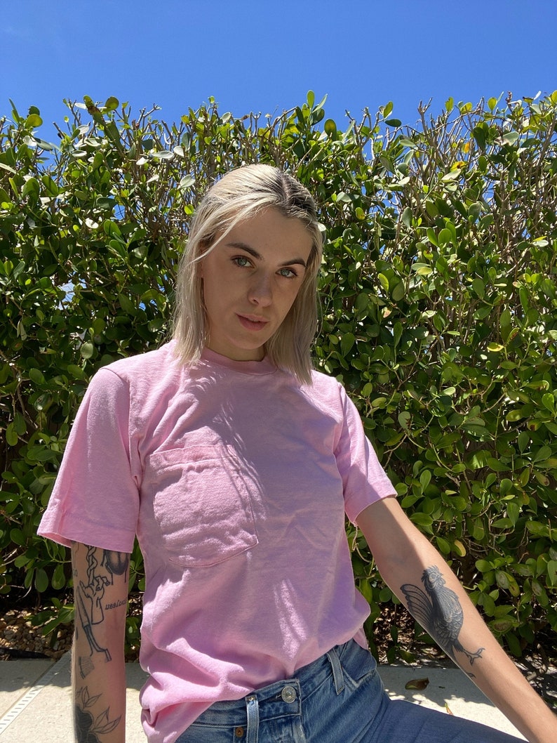 May include: A woman wearing a pink t-shirt with a pocket and blue jeans. She has short blonde hair and tattoos on her arms.