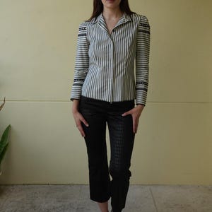 Pode incluir: Uma mulher vestindo uma camisa listrada e cal&ccedil;as pretas. A camisa tem listras verticais em tons de cinza e branco, com detalhes pretos nos ombros e punhos. As cal&ccedil;as s&atilde;o pretas com um padr&atilde;o sutil.