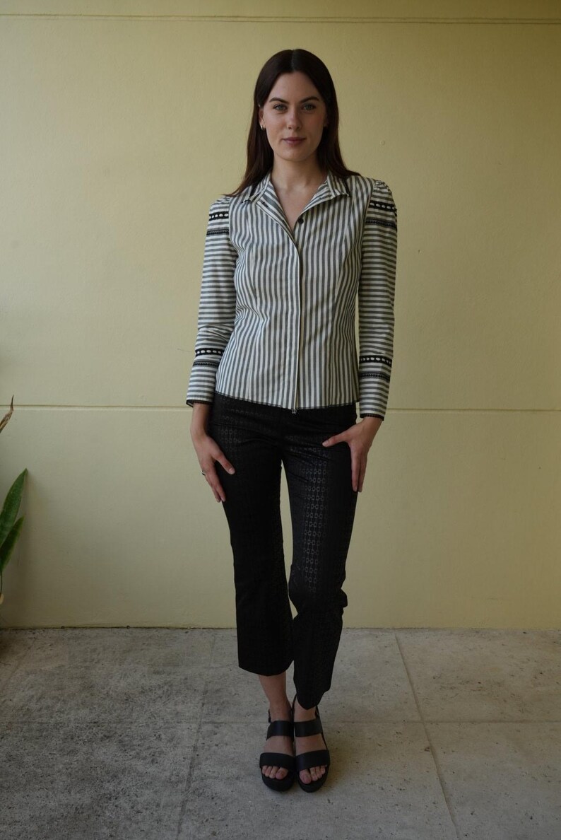 Pode incluir: Uma mulher vestindo uma camisa listrada e cal&ccedil;as pretas. A camisa tem listras verticais em tons de cinza e branco, com detalhes pretos nos ombros e punhos. As cal&ccedil;as s&atilde;o pretas com um padr&atilde;o sutil.